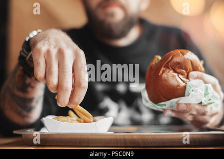 Jeune homme barbu eating hamburger et frites. Banque D'Images