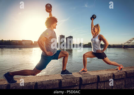 Jeune couple doing fitness entraînement avec haltères courtes sur le mur par la rivière dans un coucher du soleil. Banque D'Images