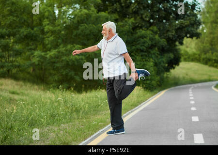 Sportif Senior faisant les exercices d'étirement sur l'hippodrome de la ville. Garder corps en forme, musclé, solide. Le maintien d'un mode de vie sain. Homme portant un polo blanc, un pantalon noir, des chaussures de sport. Positif. Banque D'Images