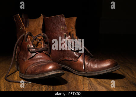 Gros plan sur des bottines en cuir marron traditionnel pour hommes, lacets défaits, sur un parquet avec fond sombre.Éclairage artistique à faible intensité. Banque D'Images