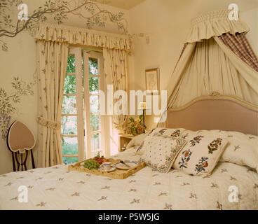 Coronet avec rideaux crème au-dessus de lit avec des coussins de fleurs dans la chambre avec mur de fleurs au pochoir et pendant à rideaux sur des fenêtres à la française Banque D'Images