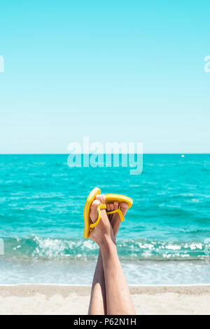 Gros plan des jambes d'un jeune homme de race blanche sur la plage, à l'envers, portant une paire de tongs jaune, contre le ciel avec un espace vide sur le dessus Banque D'Images