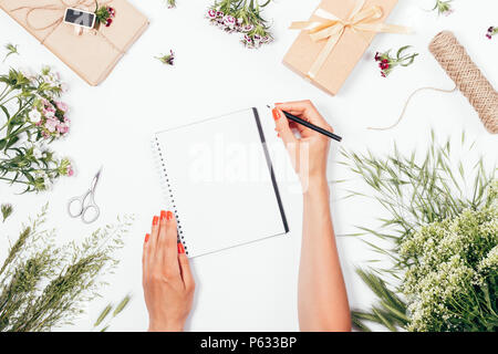 Femme de prendre des notes de l'été festif entre Fleurs et cadeaux, vue du dessus. Composition florale mise à plat avec des mains écrit journal. Banque D'Images