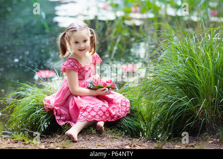 Enfant assis au bord du lac de l'eau regardant lily fleurs. Little girl holding lily rose fleur. Enfant regardant à nénuphars aux beaux jours du printemps. Piscine pour enfants Banque D'Images