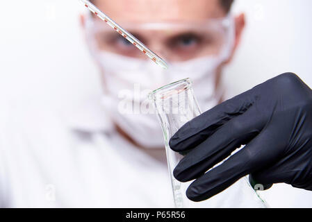 Un tube à essai et une ampoule de verre dans les mains d'un scientifique, portant des lunettes, un masque et des gants noirs sur fond blanc. Banque D'Images