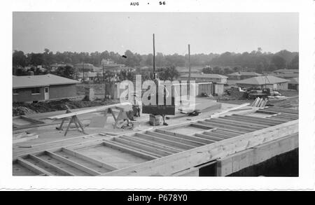 Photographie noir et blanc, montrant une femme blonde tenant un panneau en bois avec les lettres 'américain', et un petit garçon, portant un chapeau de cowboy, position assise et debout au milieu d'un site de construction, peut-être sur un toit, avec l'ossature en bois visible dans l'avant-plan, et la saleté, les débris de construction et maisons en arrière-plan, probablement photographié dans l'Ohio, Août, 1956. () Banque D'Images