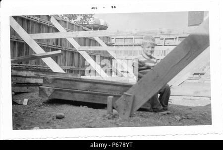 Photographie noir et blanc, montrant un petit garçon aux cheveux blonds, portant une chemise rayée, et souriant à la caméra en position assise sur plusieurs longueurs de bois en appui sur le sol de terre, entourée d'éléments de charpente, avec un château d'eau visible à l'arrière-plan, probablement photographié dans l'Ohio, Août, 1956. () Banque D'Images