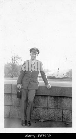 Photographie noir et blanc, montrant un soldat de sexe masculin, en pleine longueur, face à la caméra et souriant, portant un uniforme et capuchon, tenant un appareil photo, et s'appuyant sur un muret en pierre avec un parc et des monuments dans l'arrière-plan, probablement photographié dans l'Ohio pendant la Seconde Guerre mondiale, 1945. () Banque D'Images