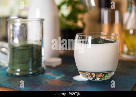 Vue rapprochée de kéfir frais probiotik verre en verre clair et vert avec une cuillère de poudre de spiruline prêt à mélanger sur une table de cuisine. Banque D'Images