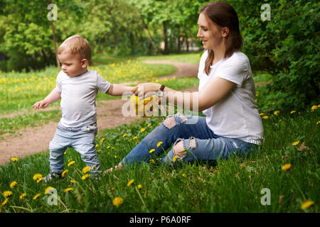 Jeune femme enceinte se trouve sur pelouse au parc de la ville. Girl détient bouquet de fleurs sur fond jaune des pissenlits en ses mains et tient son fils par la main. Banque D'Images