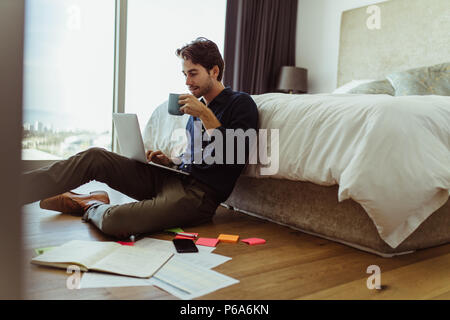 homme d'affaires travaillant sur ordinateur portable assis à la maison tout en buvant du café. Homme travaillant sur un ordinateur portable assis sur le sol avec son journal et o Banque D'Images