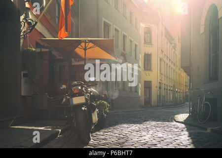 Des motos, des vélos à un café de la rue sous un parasol sur la rue avec cobblestone dans la douce lumière du soir. Riga, Lettonie Banque D'Images