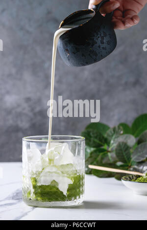 Verser la crème de pichet à thé vert matcha latte glacé ou un cocktail dans le verre, avec des cubes de glace, poudre matcha sur table de marbre blanc, décorée par green br Banque D'Images