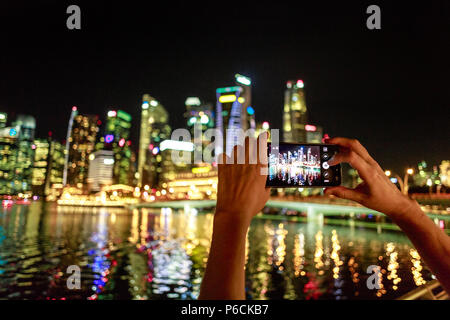 View of woman's hands prendre photo sur téléphone mobile appareil photo de Singapour marina et du quartier financier de la ville. Touchant et en maintenant la main mobile pour capturer une photo.Nuit un arrière-plan lumineux Banque D'Images