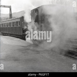 Années 1950, la vapeur envahit le conducteur dans sa cabine, comme il l'apprécier une cigarette que la petite locomotive touristique attend sur la plate-forme à l'Snowdon Mountain Railway, le Pays de Galles, Royaume-Uni. Banque D'Images