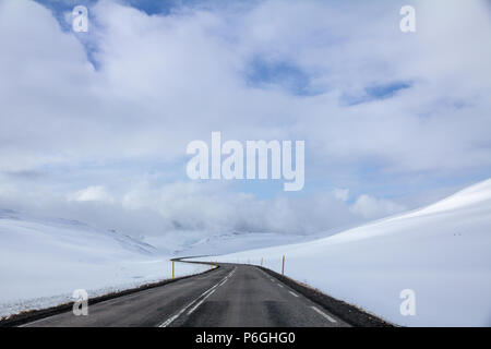 Après un printemps tardif de neige dans le nord de l'Islande, la route serpente vers l'ouest à travers le cercle de collines couvertes de neige sous un ciel partiellement nuageux. Banque D'Images