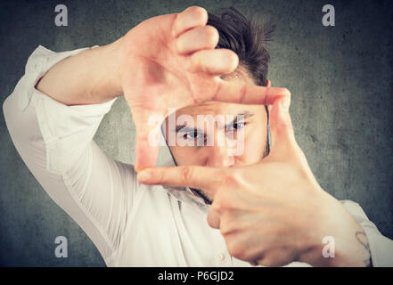 Jeune homme de faire un cadre avec les mains et les doigts à la recherche à travers les limites à appareil photo sur fond gris Banque D'Images