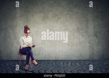 Femme élégante exerçant son activité et l'écriture sur le presse-papiers à remplir pour reprendre l'entrevue d'emploi tout en étant assis sur une chaise Banque D'Images