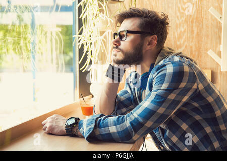 Vue latérale du jeune homme à lunettes hippie assise seule dans une cafétéria à l'écart dans la fenêtre dans la tristesse Banque D'Images