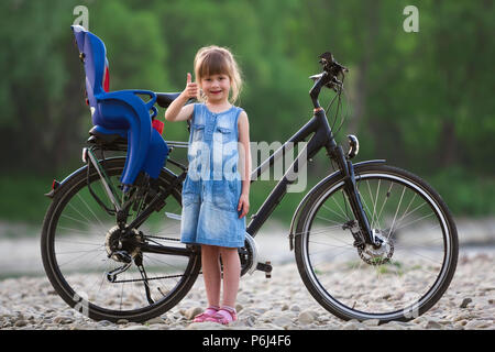 Petit très jolie jeune femme en robe bleue faire Thumbs up in front of modern vélo avec siège enfant aux arbres verts brouillée sur fond de l'été Banque D'Images