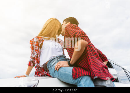 Couple assis sur le capot de leur voiture. Belle jeune femme embrasse l'homme pour le cou. L'homme, l'embrasse. close up par la rivière Banque D'Images