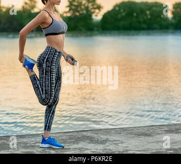 Photo recadrée de girl making exercices sur l'air frais. Les étirements, yoga, remise en forme. Le maintien du modèle de vie sain, la figure sportive, belle apparence. Lac de l'arrière-plan. Soir, belle nature. Banque D'Images