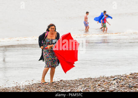 La ville de Sidmouth, Devon, UK. 1er juillet 2018. Météo britannique. Une femme sur la plage à la station balnéaire de Sidmouth dans le Devon avec parapluie, ce qui est pris par une rafale de vent qui tourne à l'envers comme la ville attrape le bord d'une adoption forte pluie douche. Crédit photo : Graham Hunt/Alamy Live News Banque D'Images