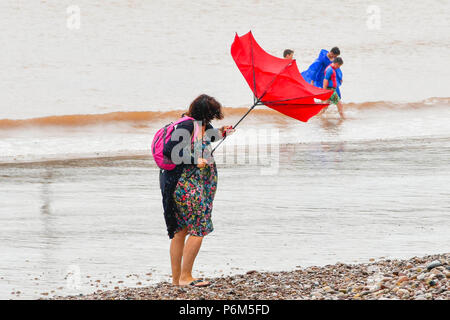 La ville de Sidmouth, Devon, UK. 1er juillet 2018. Météo britannique. Une femme sur la plage à la station balnéaire de Sidmouth dans le Devon avec parapluie, ce qui est pris par une rafale de vent qui tourne à l'envers comme la ville attrape le bord d'une adoption forte pluie douche. Crédit photo : Graham Hunt/Alamy Live News Banque D'Images