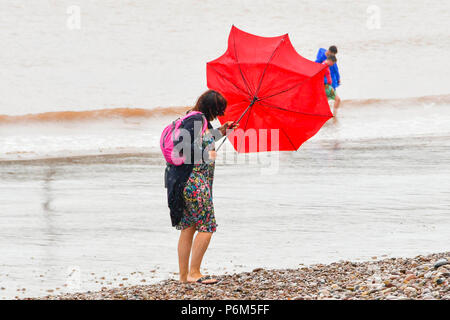 La ville de Sidmouth, Devon, UK. 1er juillet 2018. Météo britannique. Une femme sur la plage à la station balnéaire de Sidmouth dans le Devon avec parapluie, ce qui est pris par une rafale de vent qui tourne à l'envers comme la ville attrape le bord d'une adoption forte pluie douche. Crédit photo : Graham Hunt/Alamy Live News Banque D'Images