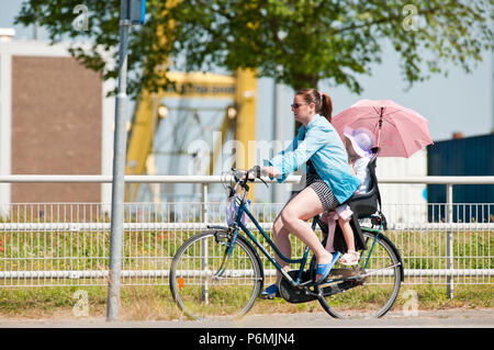 Jeune femme est à cheval sur le vélo avec un petit enfant à l'arrière, sur le dessus de l'enfant d'un parasol pour se protéger contre le soleil Banque D'Images