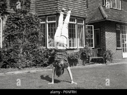 Années 1960, jeune fille faisant un ATR en extérieur dans un jardin d'une maison. Elle a ses pieds dans l'air comme elle équilibre ses mains sur le sol. Banque D'Images