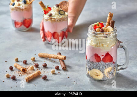 A woman's hand est titulaire d'une vitre avec un dessert frais sain composé de yaourts, de crème, de chia bio de graines, de fruits et de tubules wafer sur un béton gris, blanc Banque D'Images