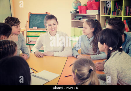 Smiling female teacher positif parler aux enfants, assis ensemble autour de desk in classroom Banque D'Images