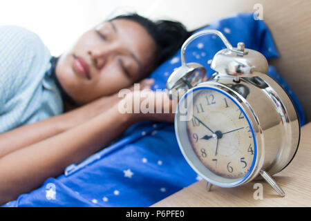 Jeune femme en robe de nuit de sommeil. La jeune fille est en train de dormir dans le lit avec un réveil à côté du lit. Banque D'Images