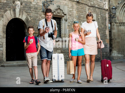 Jeune famille de quatre avec des valises à marcher le long en utilisant un téléphone Banque D'Images