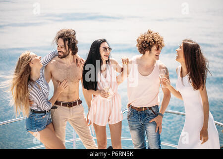 Vacances de luxe. Les jeunes joyeux amis masculins et féminins posant sur un arc de pont de bateau à voile, souriant, serrant sur fond marin bleu incroyable. Bann Banque D'Images