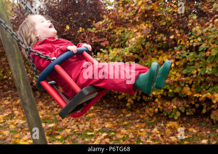 Petite fille portant un costume vert et rouge flaque wellies assis sur une balançoire Banque D'Images