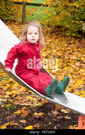 Petite fille portant un costume vert et rouge flaque wellies assis sur une diapositive Banque D'Images