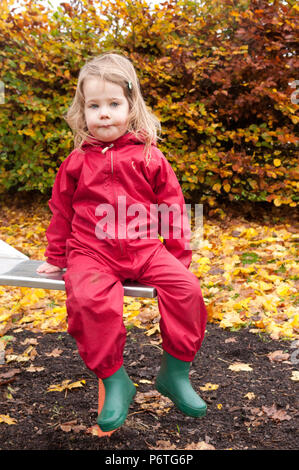 Petite fille portant un costume vert et rouge flaque wellies assis sur une diapositive Banque D'Images