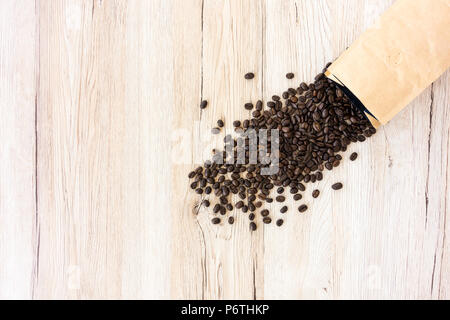 Les grains de café torréfié foncé épars et un sac en papier blanc brun rempli de grains de café sur fond de bois brun clair avec beaucoup d'espace de copie. Banque D'Images