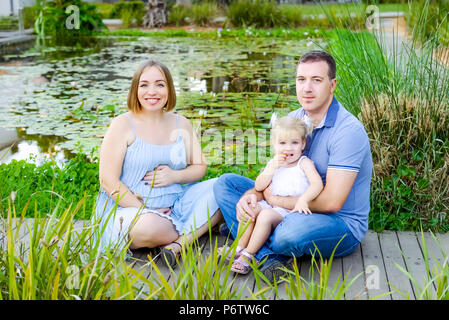 Une famille heureuse de trois - femme enceinte, père et fille s'amusant près de l'étang de nénuphars dans le parc. Loisirs en famille, passer du temps ensemble Banque D'Images