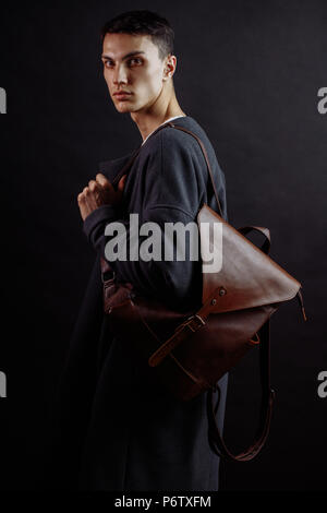 Young businessman looking at camera with serious expression vêtu d'un pardessus tendance portant sur l'épaule Sac en cuir brun. Le meilleur mode de vie, Banque D'Images