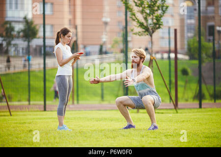 Le travail d'équipe dans les sports. Une femelle est formateur de la formation d'un jeune homme séduisant, avec une barbe dans un parc sur l'herbe verte, une pelouse au coucher du soleil. Une fille est titulaire d'un téléphone dans sa main et utilise un chronomètre Banque D'Images