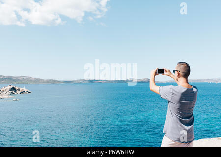 Libre d'un jeune homme de race blanche vue de derrière en prenant une photo de la mer sur la Costa Smeralda, en Sardaigne, Italie, avec son smartphone, avec une bla Banque D'Images