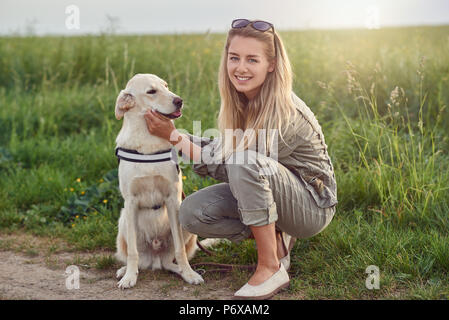 Happy smiling golden dog portant un faisceau marche assis face à sa jolie jeune femme propriétaire qui est caressant d'amour avec un sourire à l'extérieur dans cou Banque D'Images