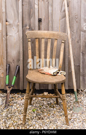 Une vieille chaise en bois avec des gants de travail et outils de jardin appuyé contre un hangar en bois idéal pour la retraite ou les concepts de jardinage Banque D'Images