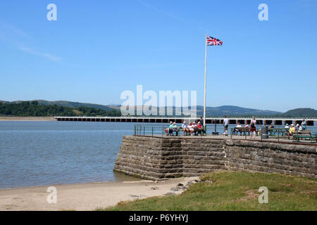 Les gens sur le quai à Arnside par le fleuve de l'estuaire de Kent à marée montante avec Arnside viaduc de chemin de fer derrière le drapeau de l'Union et sur le mât. Banque D'Images