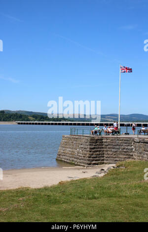 Les gens sur le quai à Arnside par le fleuve de l'estuaire de Kent à marée montante avec Arnside viaduc de chemin de fer derrière le drapeau de l'Union et sur le mât. Banque D'Images