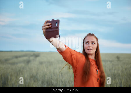 Belle jeune femme en robe rouge et les cheveux rouges, prend un au téléphone selfies dans le champ de blé vert le soir au coucher du soleil en été. la nature de la technologie numérique. Banque D'Images