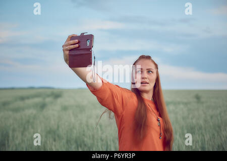 Belle jeune femme en robe rouge et les cheveux rouges, prend un au téléphone selfies dans le champ de blé vert le soir au coucher du soleil en été. la nature de la technologie numérique. Banque D'Images